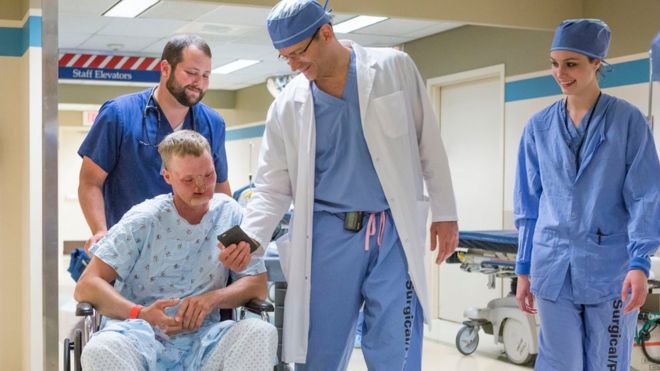Доктор Самир Мардани показывает Энди, который находится в инвалидной коляске и в больничном халате, фотографии своих детей за мгновение до того, как они отправятся в операцию. Оба улыбаются.