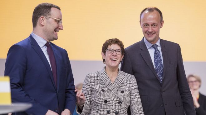 Аннегрет Крамп-Карренбауэр стоит, получив большинство голосов, чтобы стать следующим лидером немецких христианских демократов (ХДС) между Йенсом Спаном (слева) и Фридрихом Мерцем (справа)