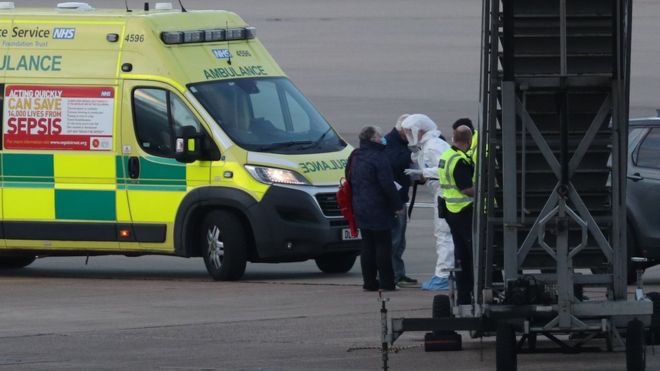 Член Служба скорой помощи NHS West Midlands, ношение защитной одежды и маска для лица разговаривает с пассажирами (слева) круизного лайнера Grand Princess, пострадавшего от коронавируса, после того, как они прибыли в аэропорт Бирмингема после репатриации в Великобританию из США