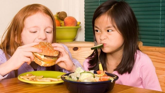 Girl quietly eating cucumber get annoyed as her friend makes noises while eating a giant hamburger