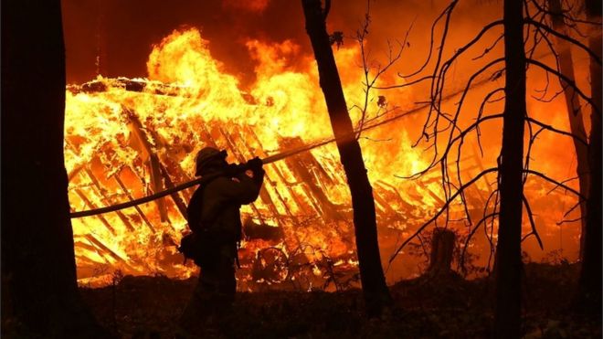 Пожарный Cal Fire разбрызгивает воду на дом рядом с горящим домом, когда пожарный лагерь движется по территории