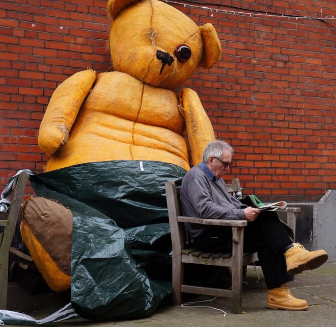 A man reads in front of a large teddy bear