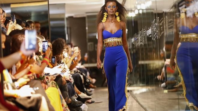 A model walking down a catwalk during African Fashion International Fashion Week in Cape Town, South Africa as people in the crowd take pictures. The model is wearing clothes by UNICEF ambassador and South African designer Gavin Rajah.