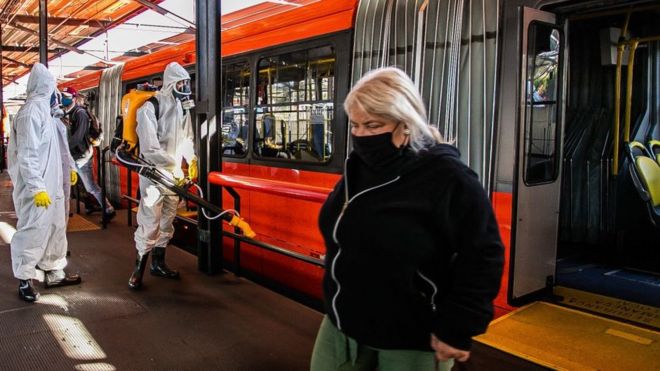 Mulher em ponto de ônibus em Curitiba com profissionais desinfectando o local