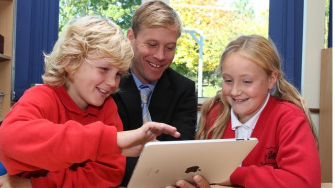 Professor (ao centro) recebe ensinamentos de um garoto (esq.) e uma menina (dir.) em um tablet