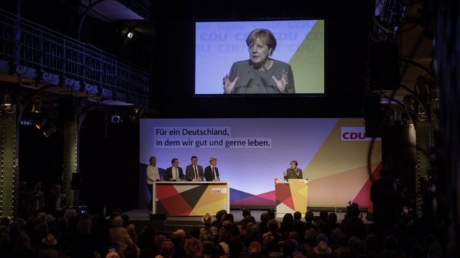 Политики ХДС наблюдают за Меркель на сцене перед аудиторией в закрытом зале