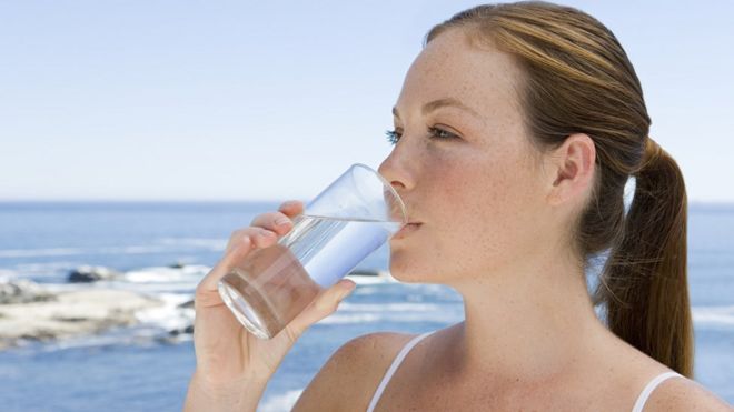 Mujer bebe agua en un vaso con el mar de fondo.