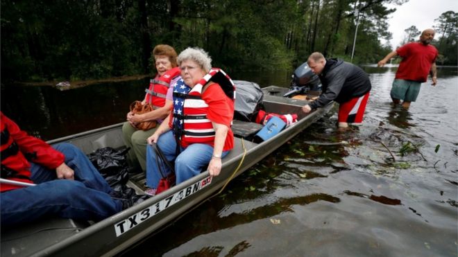 Imagem mostra homens de pé no chão e duas mulheres sendo resgatadas em embarcação em área inundada após a passagem do furacão Florence