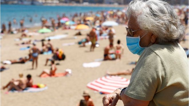 Женщина в маске наблюдает за людьми, загорающими на пляже Лас-Кантерас, поскольку некоторым испанским провинциям разрешено ослабить ограничения на изоляцию во время второй фазы на фоне коронавирусной болезни