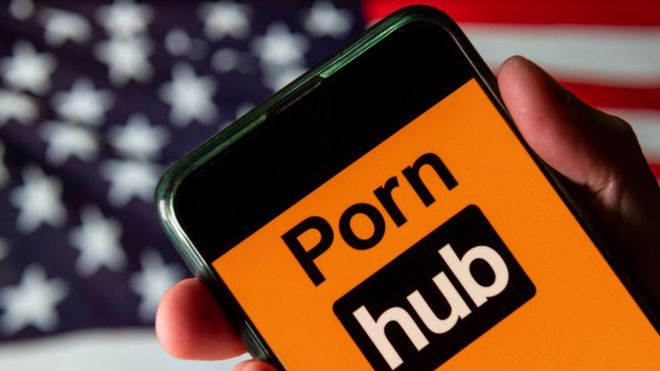 The Pornhub logo is seen on a mobile phone screen held by a hand in front of an American flag