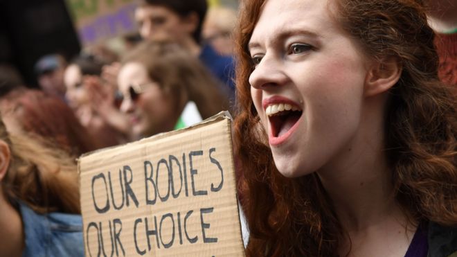 A woman, open mouthed, at a rally holds up a sign saying 'our bodies, our choice'