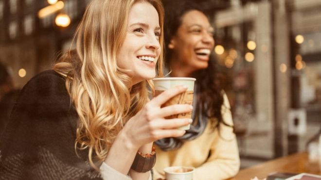 Фото показывая 2 молодых женщины смеясь над и выпивая кофе в кофейне.