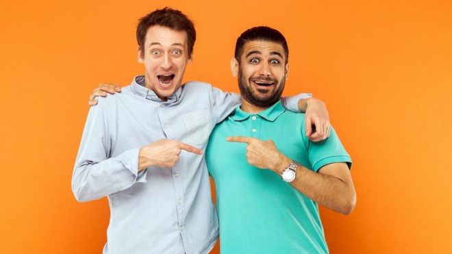 Two best friends hugging, pointing finger each other and looking at camera with shocked face - orange background