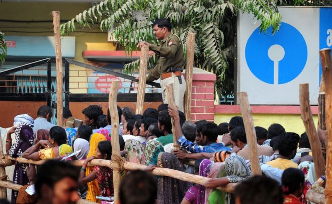 Индийский полицейский контролирует толпу, поскольку жители деревень выстраиваются в очередь возле банка, ожидая, чтобы 18 ноября 2016 года внести депозит и обменять 500 и 1000 рупийных банкнот в деревне Хануман Гандж на окраине Аллахабада.