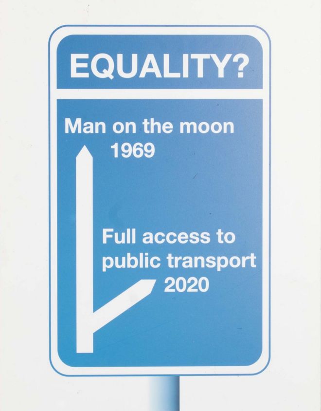 Синий дорожный знак с двумя стрелками. В верхней части написано «Человек на луне 1969», в нижней - «Полный доступ к общественному транспорту 2020».
