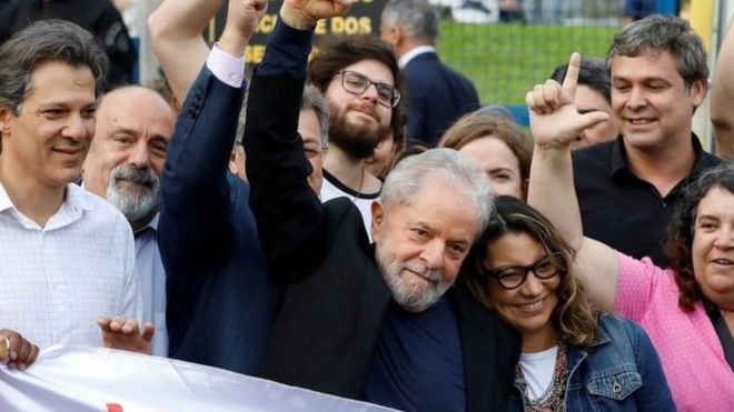 Lula cercado de apoiadores faz gesto com os dedos imitando a letra L