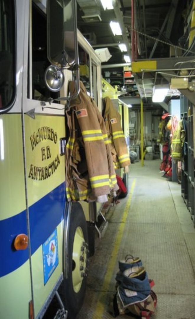 Пожарная машина читает «МакМердо Ф.Д. Антарктида. Он припаркован в пожарной части, рядом с ним хранится противопожарное снаряжение и оборудование