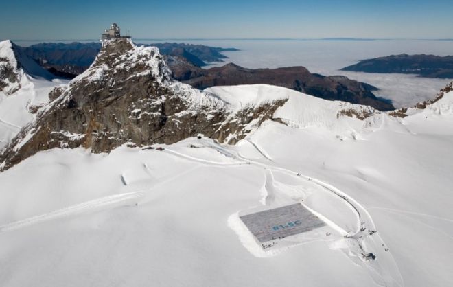 Увеличенный вид с воздуха показывает гигантскую открытку на леднике Алеш, вокруг которой лежит снежное пространство, а за вершинами возвышаются пики