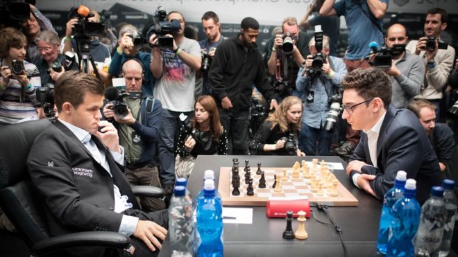 Магнус Карлсен (слева) и претендент Фабиано Каруана смотрят друг на друга за шахматной доской, в то время как множество фотографов карабкаются на заднем плане