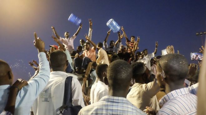Люди с большими пластиковыми контейнерами для воды аплодируют сидячим военным штабам в Хартуме, Судан - воскресенье, 7 апреля 2019 года