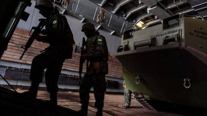 Dois soldados com uniformes do exército brasileiro seguram fuzis em frente a um tanque durante exercício militar