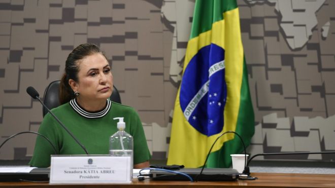 Katia Abreu com olhar sério sentada diante de mesa de comissão, com bandeira do Brasil ao fundo
