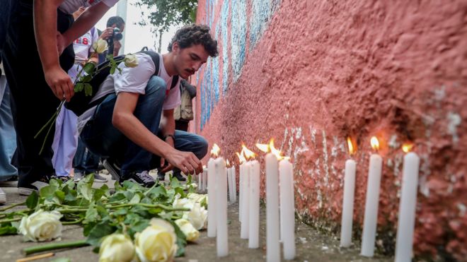 Homem branco acende vela em frente a um muro de escola, há rosas no chão