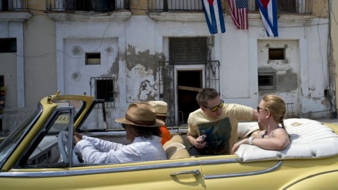 Туристы на классическом американском автомобиле проезжают мимо балкона, украшенного кубинскими и американскими флагами, в Гаване (20 марта 2016 года)