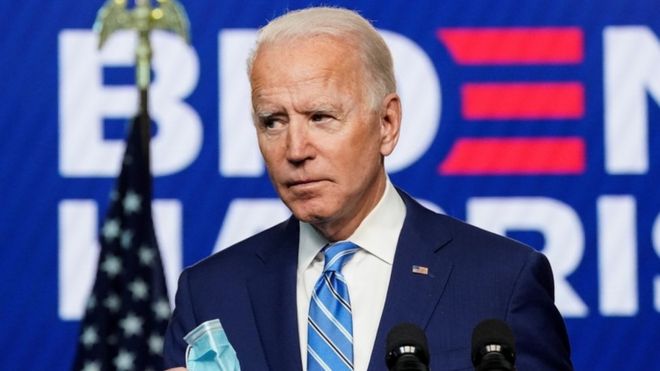Joe Biden, um homem branco de cabelos brancos, em frente a um cartaz de sua campanha eleitoral
