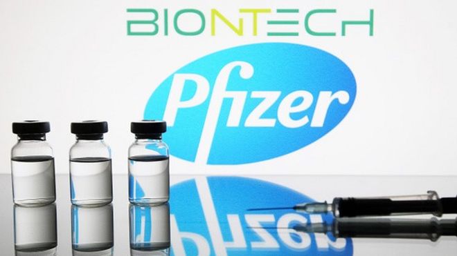 Foto de seringas e ampolas num fundo com o logo da Pfizer e da BioNTech