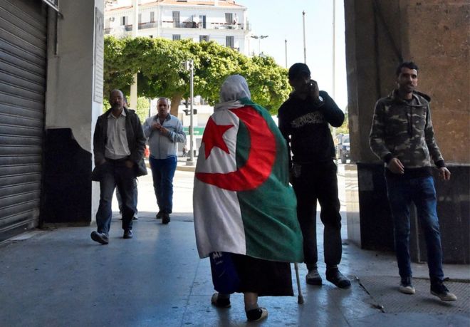 Пожилая алжирская женщина, завернутая в национальный флаг, идет по колоннаде в Алжире.