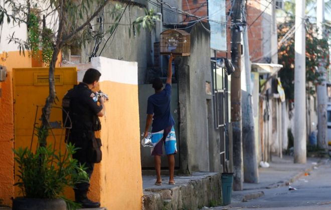 Полицейский направляет свое оружие, когда житель держит клетку для птиц во время операции против торговцев наркотиками в Сидаде-де-Деус или трущобе Города Бога в Рио-де-Жанейро