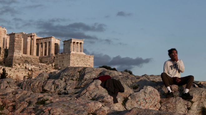 Homem sentado em colina em frente à Acrópolis de Atenas