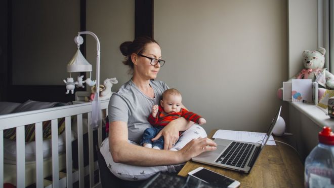 Женщина работает дома за столом в своей спальне. Она пользуется ноутбуком, держа на руках свою маленькую дочь