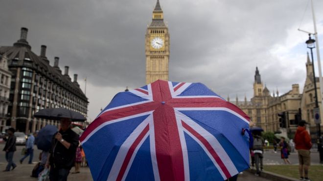 Пешеходные укрытия от дождя под зонтиком с флагом Союза, когда они идут возле циферблата Биг-Бена и башни Элизабет у здания парламента в центре Лондона 25 июня 2016 года