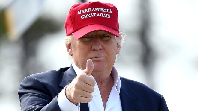 Дональд Трамп поднимает палец вверх, надевая кепку Make America Great Again