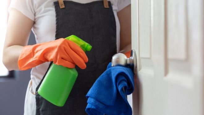 Женские руки в оранжевых резиновых перчатках очищают касающуюся поверхности двери, дверную ручку с синей салфеткой из микрофибры и зеленую бутылку с распылителем - стоковое фото