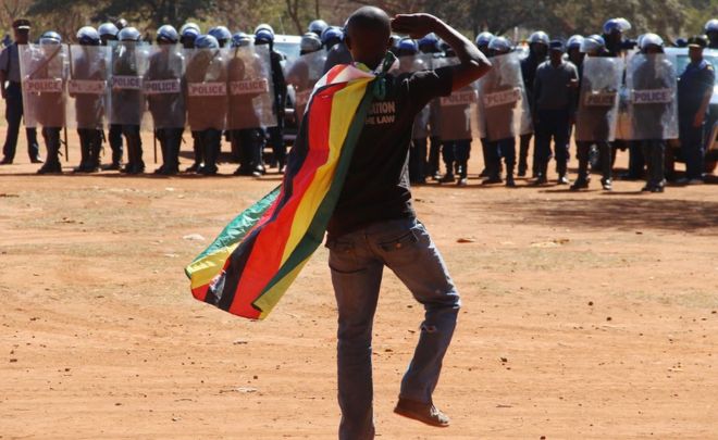 Мужчина в зимбабвийском флаге приветствует омоновцев во время акции протеста в Хараре, Зимбабве, в пятницу, 26 августа 2016 года
