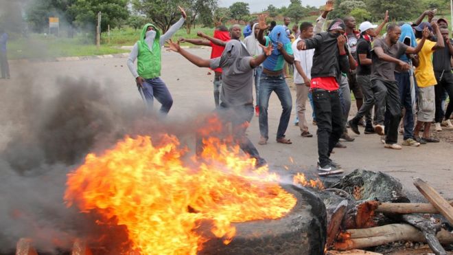 Протестующие стоят за горящей баррикадой во время акций протеста на дороге, ведущей в Хараре, Зимбабве, 15 января 2019 года.