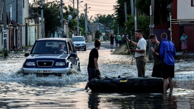 People stand next to an inflatable boat as a car drives past them in a flooded street of Kherson
