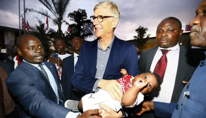 Бывший футбольный тренер Арсен Венгер (С) держит ребенка, одетого в цвета Арсенала, по прибытии в международный аэропорт Робертс в Харбеле, Либерия, 22 августа 2018 года.