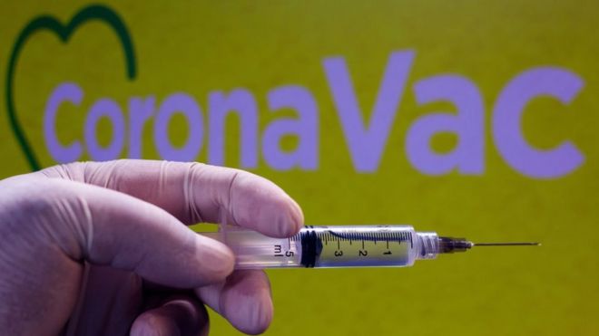 Mão com luva segura vacina diante de logo da CoronaVac
