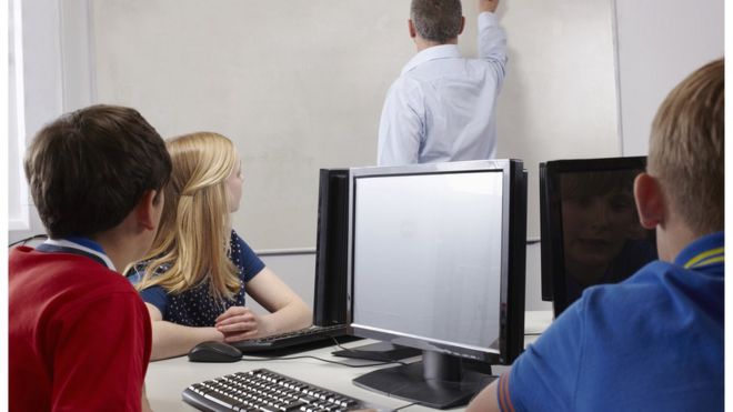 Three pupils watch a teacher while sitting at computer screens