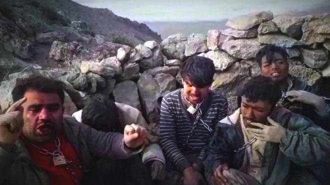 Grupo de hombres con cadenas al cuello sentados junto a un muro en la cima de una montaña