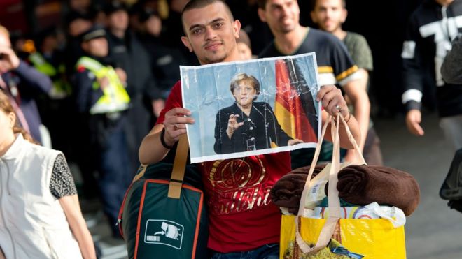 Беженец, прибывающий в Германию, держит смятую картину Ангелы Меркель