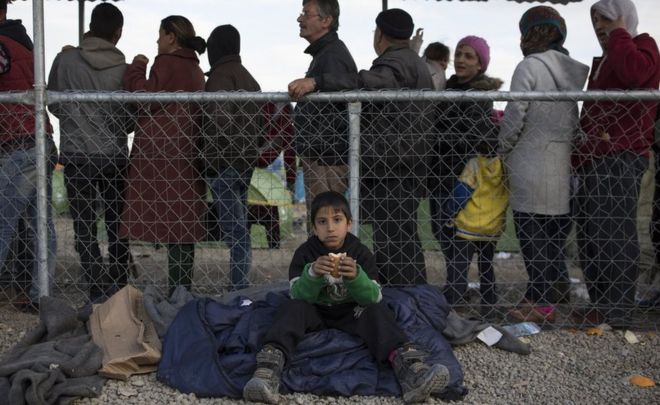 Мальчик ест бутерброд, когда люди стоят в очереди на регистрационные документы на греко-македонской границе