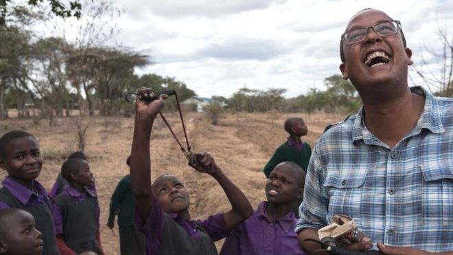 Кенийский мужчина смеется, когда дети целятся в небо катапультой