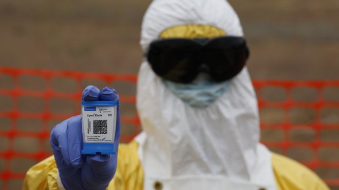 Одетый лаборант держит тестовый патрон Эбола Доктор Сенгуку из больницы Искупления в Монровии говорит, что быстрое тестирование на Эболу спасло жизни людей! Доктор Джуд Сенгуку