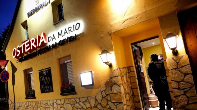 Знак на двухэтажном доме, окрашенном в желтый цвет, объявляет, что этот итальянский ресторан называется Osteria Da Mario, но на этой фотографии рассвета двое полицейских охраняют его открытую дверь