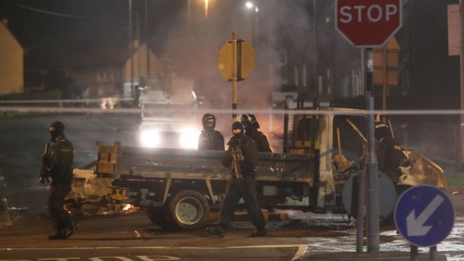 Полиция в спецназе патрулирует мимо сгоревшего автомобиля во время насилия в Лондондерри